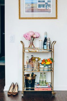 a gold bar cart with drinks and flowers on it in front of a white wall