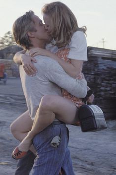 two people are hugging each other in the middle of a dirt road with stacks of wood behind them