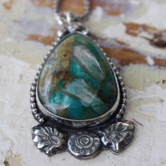 An unusual Chrysocolla teardrop-shaped stone features crystal-like matrics within the stone. The teardrop shape stone is framed with a beaded wire. At the bottom of the pendant, there are handmade Moroccan-style flower and leaf motifs giving this pendant a Moroccan flare. The cool blues and greens of this stone will pair nicely with most of your wardrobe. Moroccan Necklace, Blues And Greens, Stone Feature, Moroccan Style, The Cool, Crystals, Wardrobe, Stone, Pendant