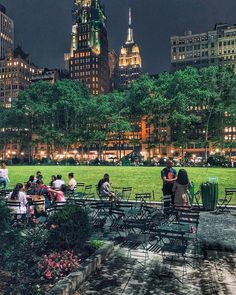 people are sitting at tables in the middle of a park with tall buildings behind them