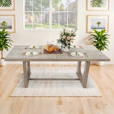 a dining table with plates and flowers on it in front of a window filled with potted plants