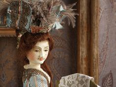 a doll with red hair wearing a blue dress and feathered headdress sits in front of a mirror