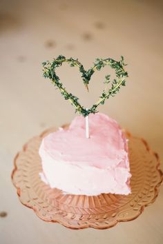 a heart shaped cake sitting on top of a pink frosted cake plate with a twig sticking out of it