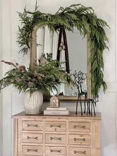 a wooden dresser topped with a white vase filled with flowers and greenery next to a mirror