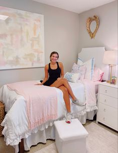 a woman sitting on top of a bed next to a white dresser and foot stool