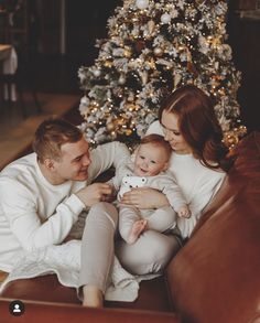 a man and woman sitting on a couch with a baby in front of a christmas tree