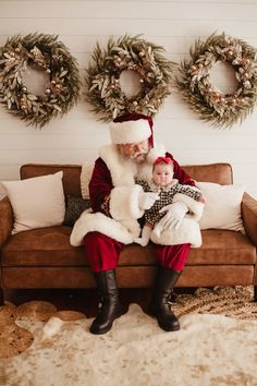 santa claus sitting on a couch with a baby in his lap and wreaths behind him