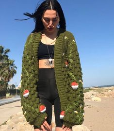 a woman standing on top of a beach next to the ocean wearing a green crocheted cardigan