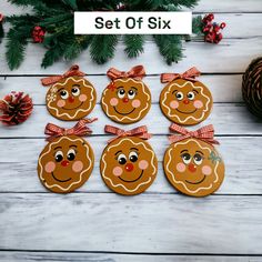 six ginger cookies with faces and bows on them sitting in front of pine cones, evergreen branches