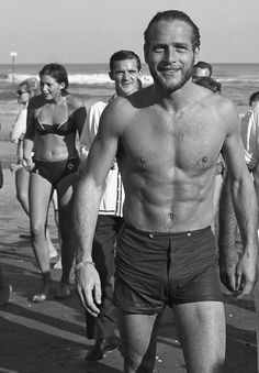 a shirtless man walking on the beach with other people in bathing suits behind him