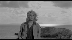 a woman standing on top of a rocky beach next to the ocean with her hands in her pockets