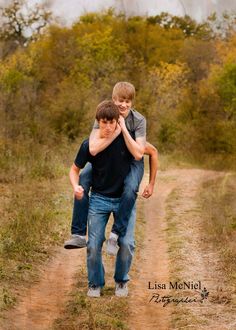 two young men walking down a dirt road in the middle of a field with trees behind them