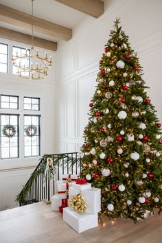 a christmas tree with presents under it in a large room next to stairs and chandelier