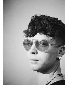 a black and white photo of a young man with glasses looking at the camera while wearing a chain around his neck