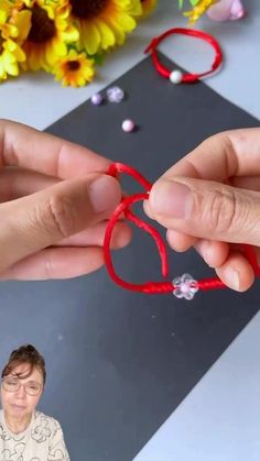 two hands holding red string with beads and flowers in the background on top of a table