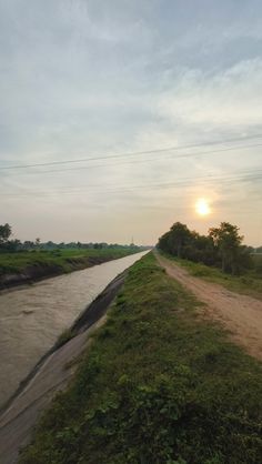 the sun is setting over a river and grassy area with dirt road in foreground