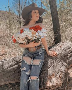 a woman wearing ripped jeans and a hat with flowers on her chest, leaning against a tree trunk