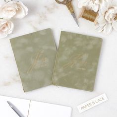 two green wedding guest book with gold writing on them and flowers next to the couple's name