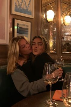 two women sitting at a table with wine glasses in front of them and one is kissing the other