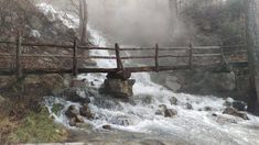 a wooden bridge over a small waterfall in the woods on a foggy winter day