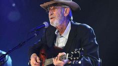 an older man wearing a hat and playing a guitar in front of a microphone on stage