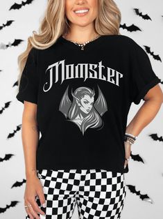 T-Shirt Description: Get in the spooky spirit with this Halloween t-shirt featuring the quote 'Momster'. This unique design showcases a stylish bat lady with sharp features, flowing hair, and bat wings against a black background, perfectly capturing the essence of the season. Stand out during the Halloween festivities with this eye-catching and hauntingly beautiful shirt that is sure to turn heads wherever you go. Embrace your inner 'Momster' and stand out this Halloween season with this unique Edgy Halloween Tops With Character Print, Edgy Halloween T-shirt With Text Print, Edgy Halloween Fan Merchandise T-shirt, Spooky Graphic Print T-shirt For Fans, Edgy Tops With Halloween Front Print, Edgy Tops With Front Print For Halloween, Edgy Halloween Tops With Text Print, Trendy Halloween T-shirt With Text Print, Trendy Halloween Text Print T-shirt