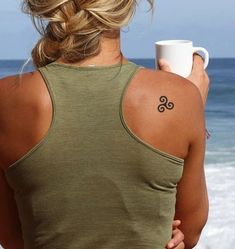 a woman with a tattoo on her back holding a coffee cup and looking at the ocean