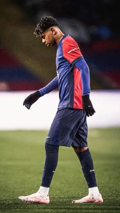 a man standing on top of a soccer field wearing a blue and red uniform with his hands in his pockets