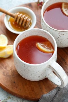 two white mugs filled with tea and honey sit on a wooden tray next to lemon slices