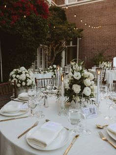 the table is set with white flowers and place settings for an elegant wedding reception in front of a brick building