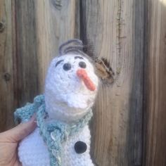 a crocheted snowman wearing a hat and scarf is posed in front of a wooden fence