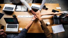 four people holding hands over a table with laptops