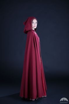 a woman in a long red dress with a hood on her head, standing against a dark background