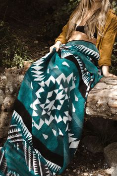 a woman is sitting on a log wearing a green and white blanket with geometric designs