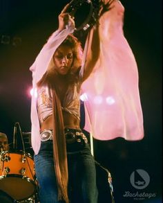 a woman with long hair standing next to a drum set on stage and holding her hands up in the air