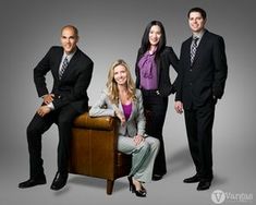 three business people posing for a photo in front of a gray background, with one woman sitting on a chair and two men standing behind them