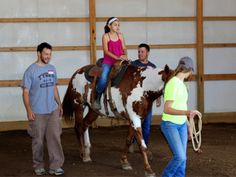 three people standing around a brown and white horse in an indoor arena with one woman sitting on top of the horse