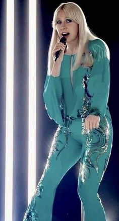 lady in blue jumpsuit singing on stage with lights behind her and microphone to the side