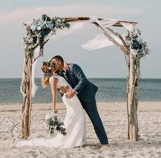 a bride and groom kissing under an arch made out of driftwood on the beach