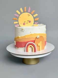 a birthday cake decorated with a sun and rainbow on top of a white plate next to a gold stand