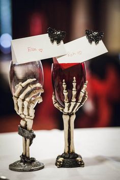 two wine glasses with skeleton hands holding each other's heads and name tags attached to them