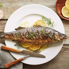 a fish is on a plate with lemons and herbs next to it, along with a knife and fork