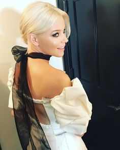 a woman with blonde hair wearing a white dress and black bow around her neck, standing in front of a door