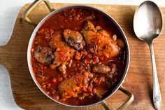 a pan filled with meat and tomato sauce on top of a wooden table next to silverware