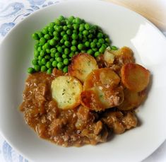 a white plate topped with meat and potatoes covered in gravy next to green peas