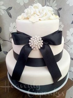 a three tiered cake with black ribbon and white flowers on the top is adorned with a crystal brooch