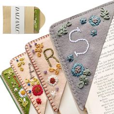 three bookmarks with embroidered letters and flowers on them next to an open book,