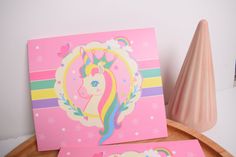 two greeting cards on a table with a pink vase in the background and a unicorn card