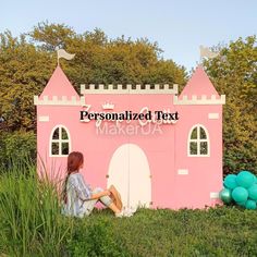 a woman sitting in front of a fake pink castle with balloons on the grass and trees behind her