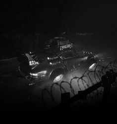 two cars in the dark with their lights on and one car is parked behind a barb wire fence